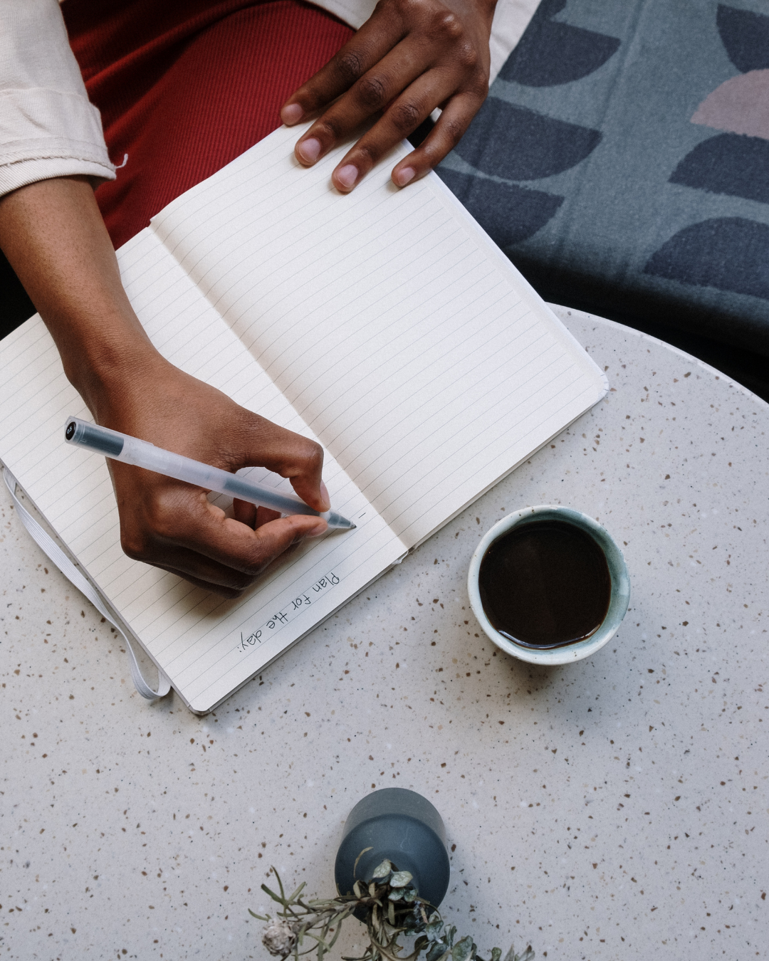 Hand writing 'Plan for the day' in a notebook beside a cup of coffee on a terrazzo table